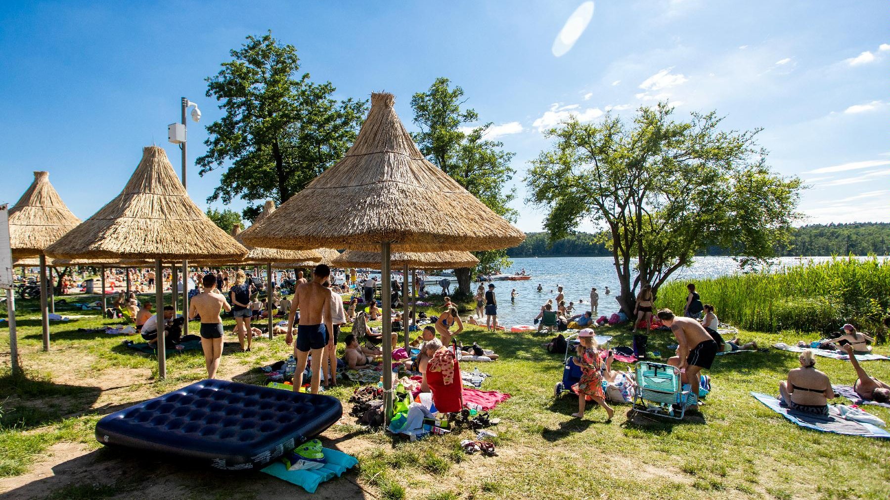 Zdjęcie przedstawia plażę - widać na nim siedzących na kocach ludzi, parasolki ze słomy oraz jezioro.