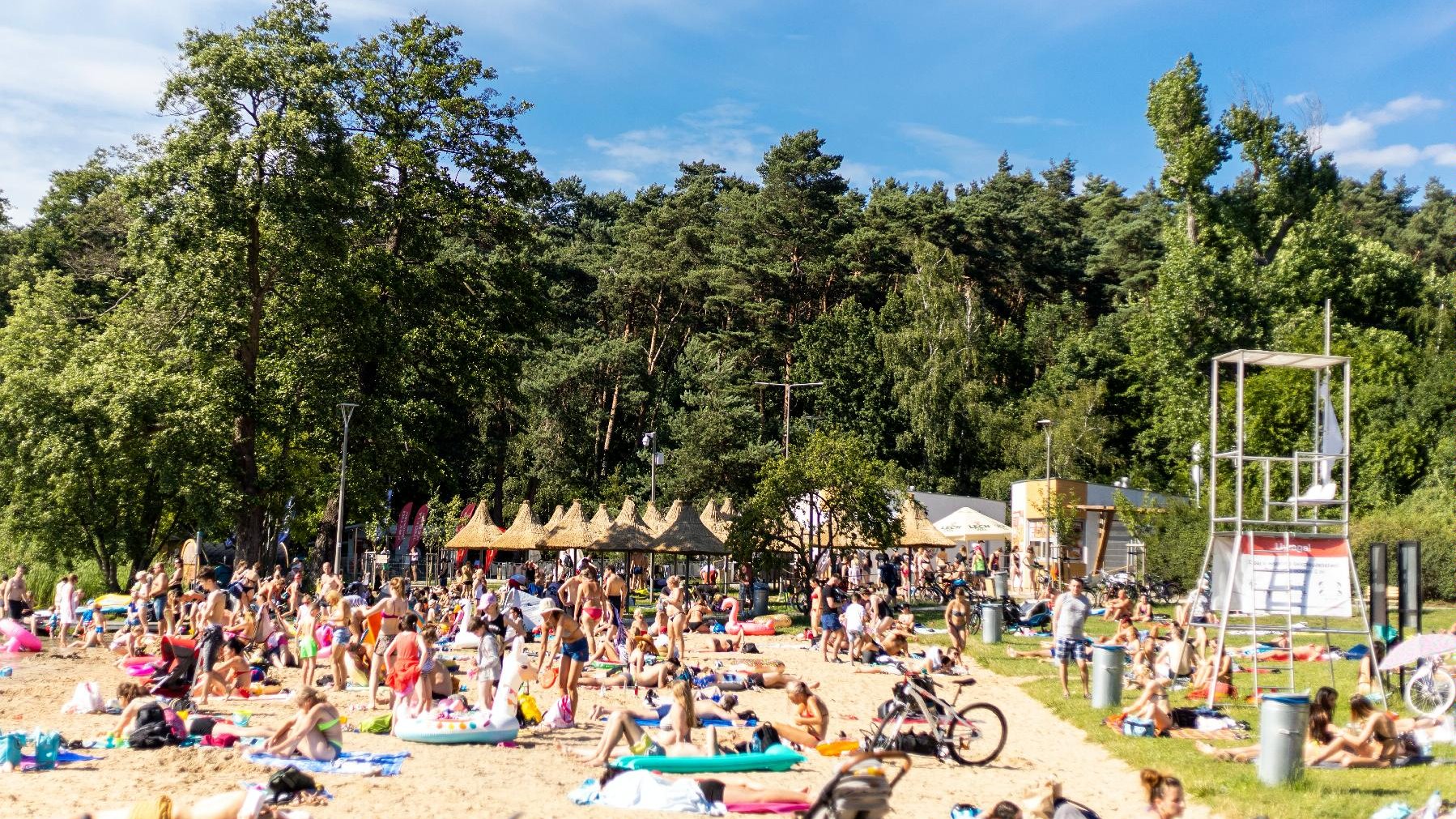 Zdjęcie przedstawia plażę - widać na nim siedzących na kocach ludzi, parasolki ze słomy, wieżę ratownika oraz zieleń.