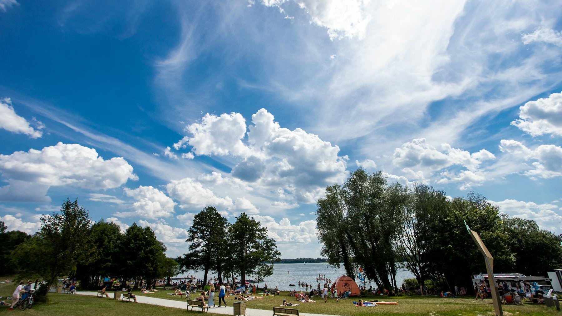 Zdjęcie przedstawia ludzi na plaży, widać na nim tereny zielone i jezioro.