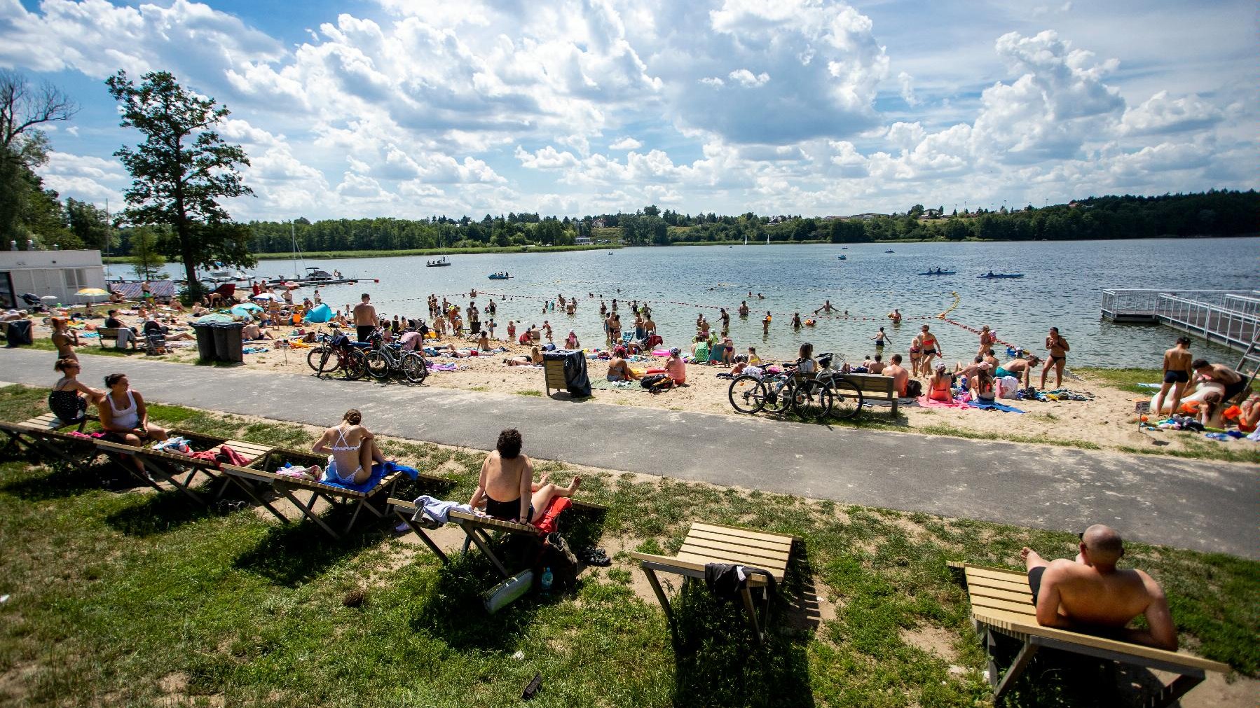 Zdjęcie przedstawia ludzi na plaży. Na pierwszym planie widać osoby na leżakach, w tle znajduje się jezioro.