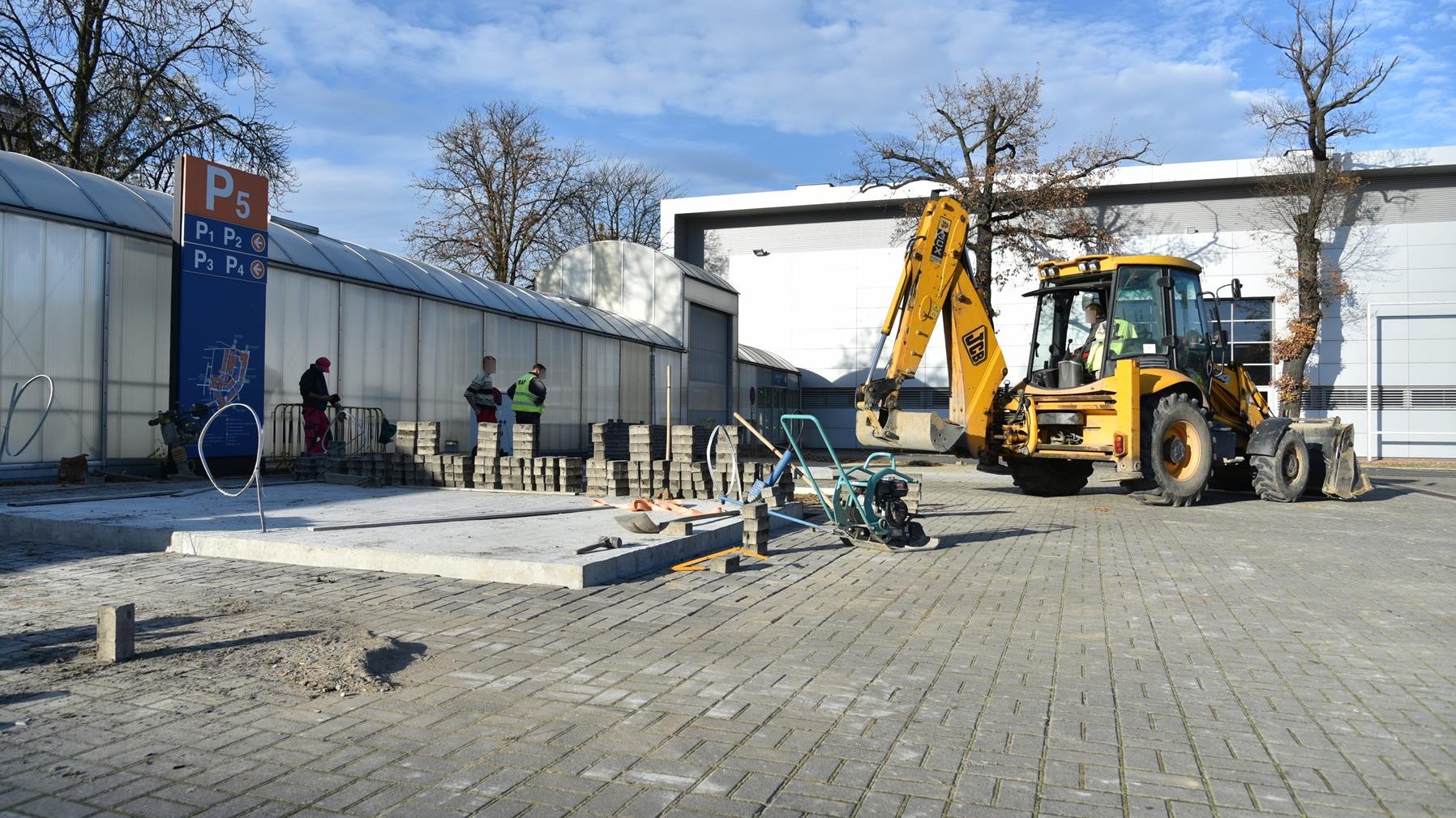 Zdjęcie: plac obok hal targowych.Na pierwszym planie świeżo wylane fundamenty i duża, żółta koparka, obok trzech mężczyzn