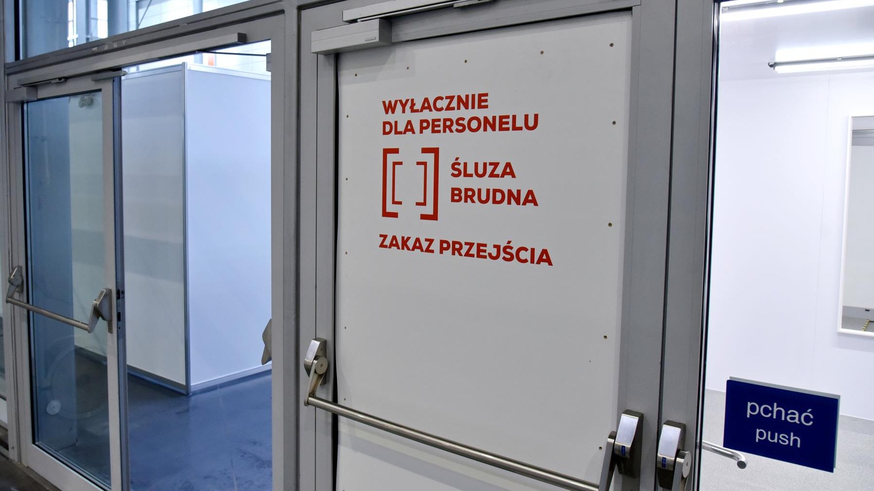 Wnętrze szpitala. Białe drzwi z czerwnonym napisem: wyłącznie dla personelu, śluza brudna, zakaz przejścia