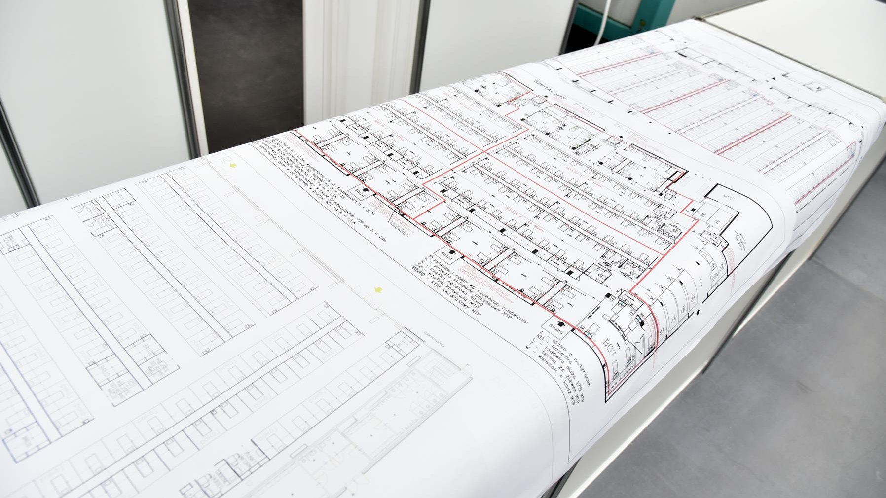 Wnętrze szpitala. Długi stół, na nim rozłożone trzy duże płachty papieru z planami szpitala