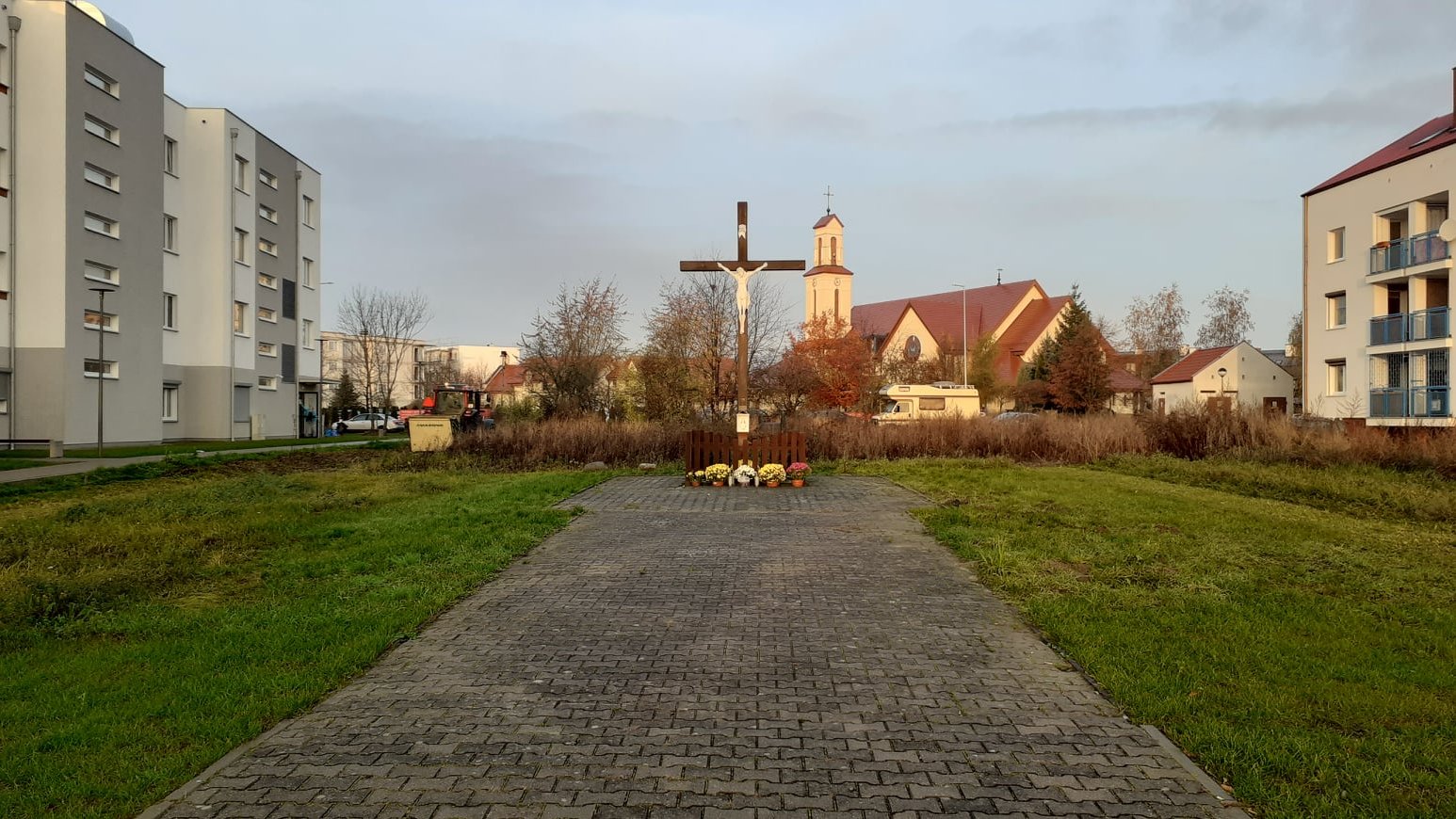 Droga, po bokach pasy zielieni. W tle krzyż.