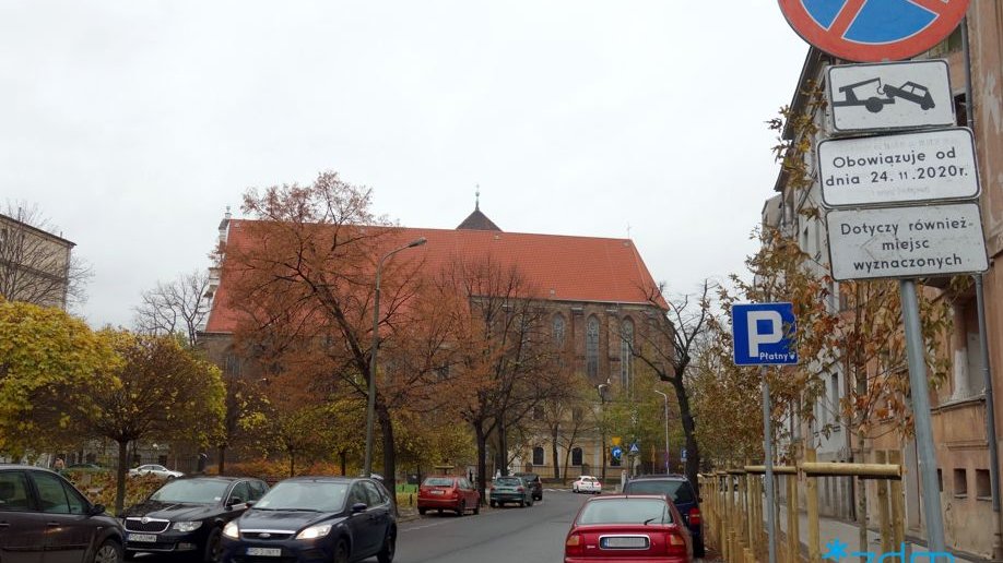 Ulica Karmelicka. Po prawej stronie znaku. Po lewej skwer. Przy ulicy parkujące samochody. W tle kościół.