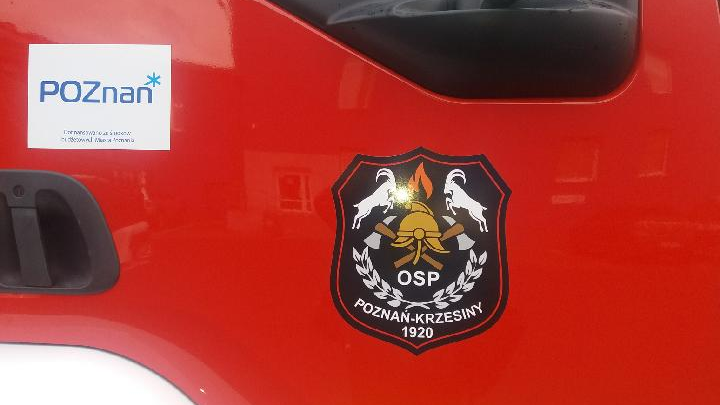 Czerwone dzwi do kabiny wozu strażackiego. Na nich naklejka "OSP Poznań-Krzesiny" oraz logo Miasta Poznania.