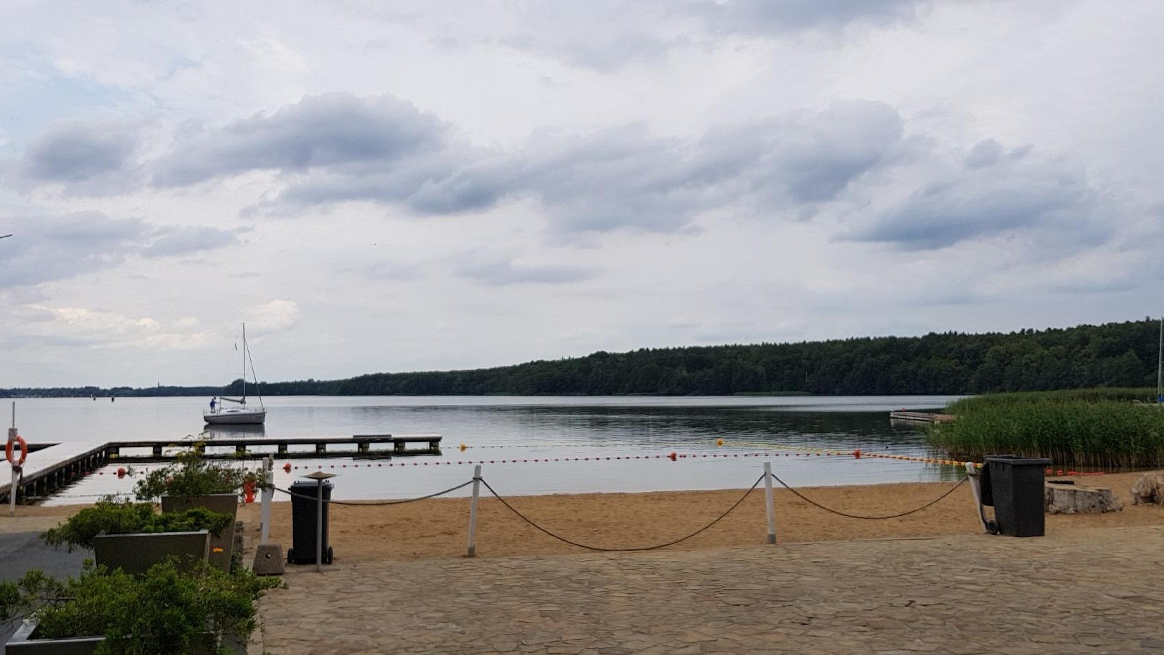 Zdjęcie przedstawia miejską plażę. Na jeziorze widać pomost i statek.