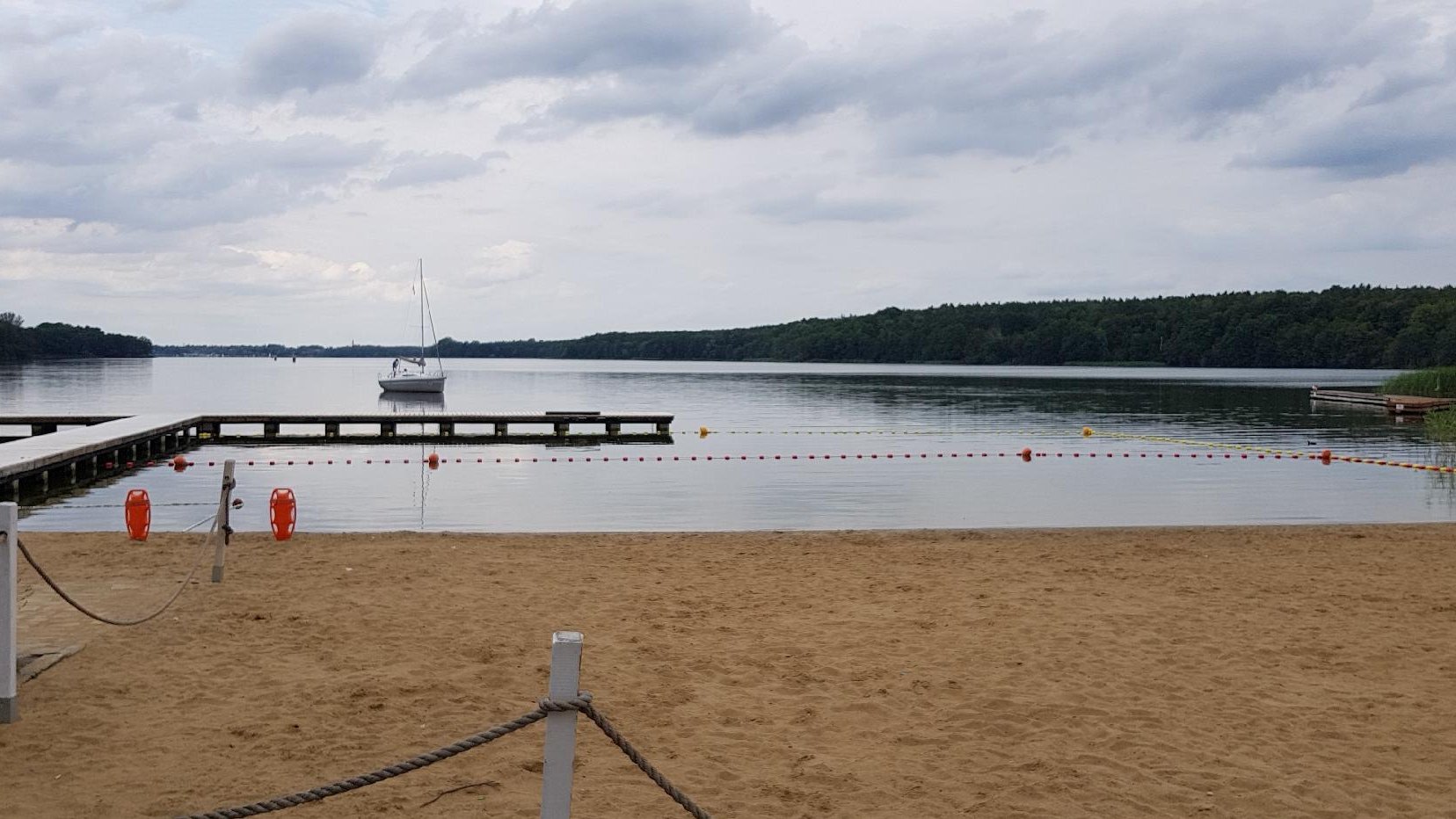 Zdjęcie przedstawia miejską plażę. Na jeziorze widać pomost i statek.