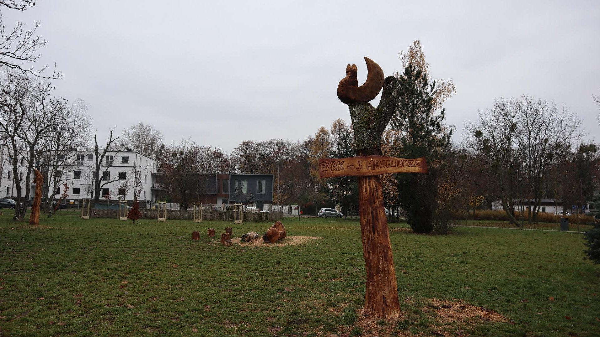 Zdjęcie przedstawia drewnianą rzeźbę z napisem "Park Heweliusza". W tle widać park.