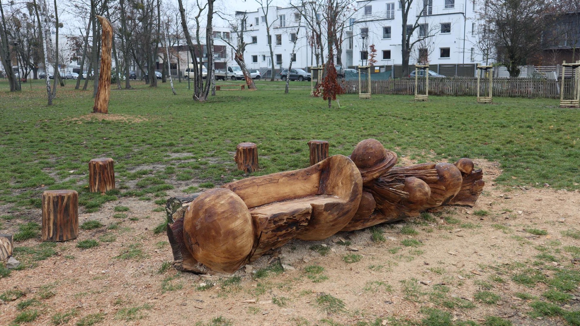 Zdjęcie przedstawia drewnianą rzeźbę pełniącą również funkcję ławki. W tle widać park.