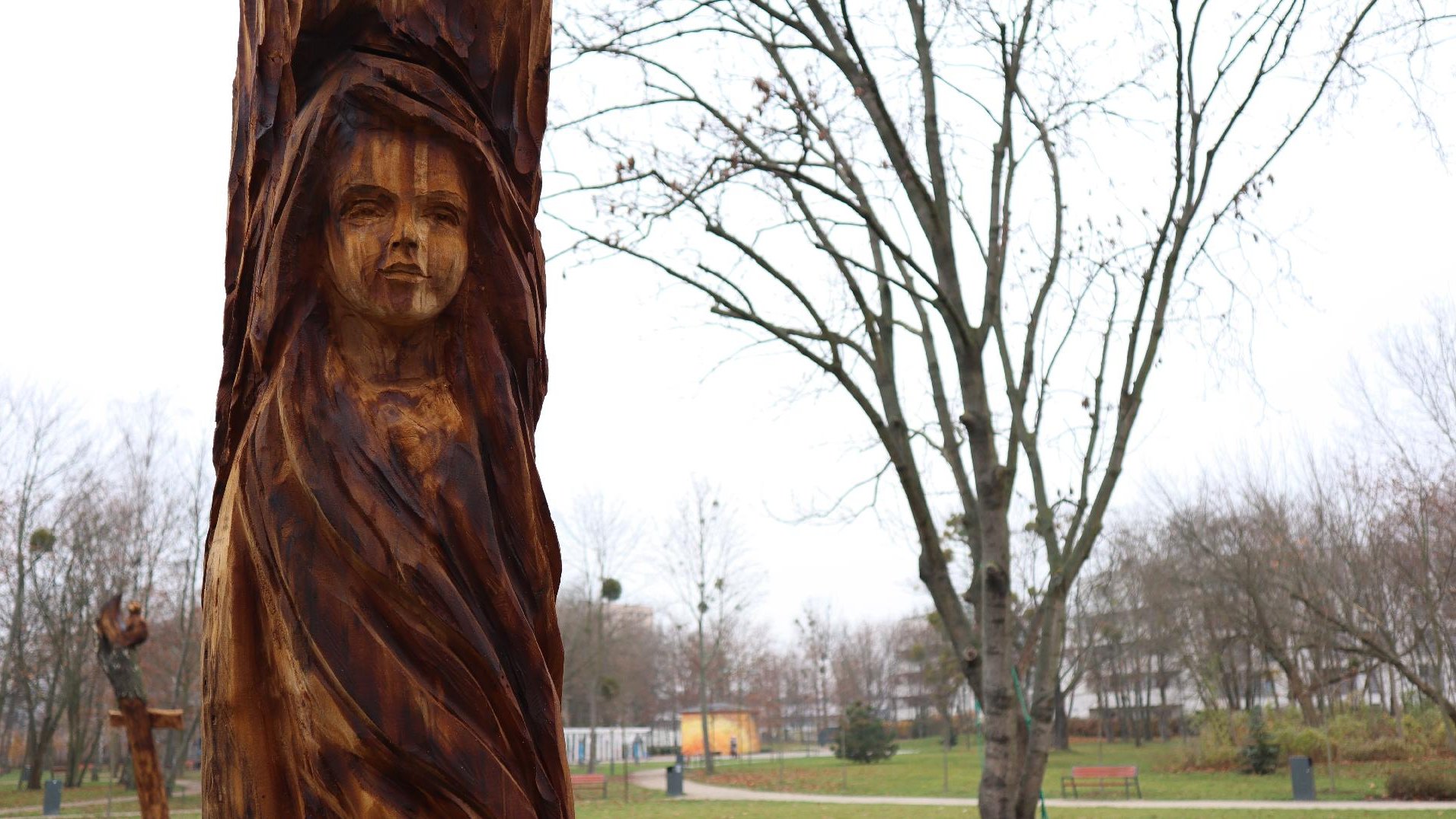 Zdjęcie przedstawia drewnianą rzeźbę dziewczyny. W tle widać park.