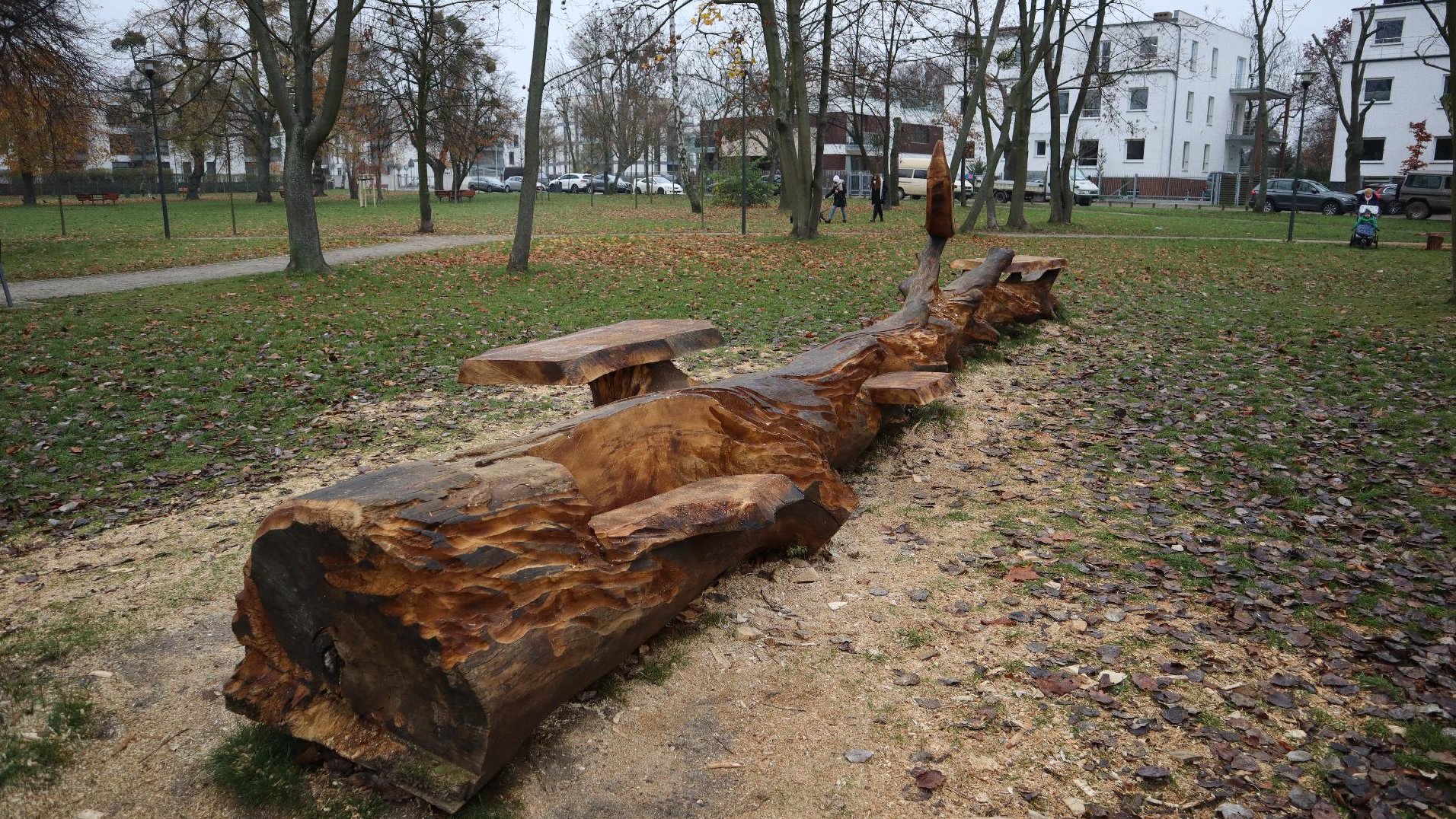 Zdjęcie przedstawia drewnianą rzeźbę pełniącą również funkcję ławki. W tle widać park.