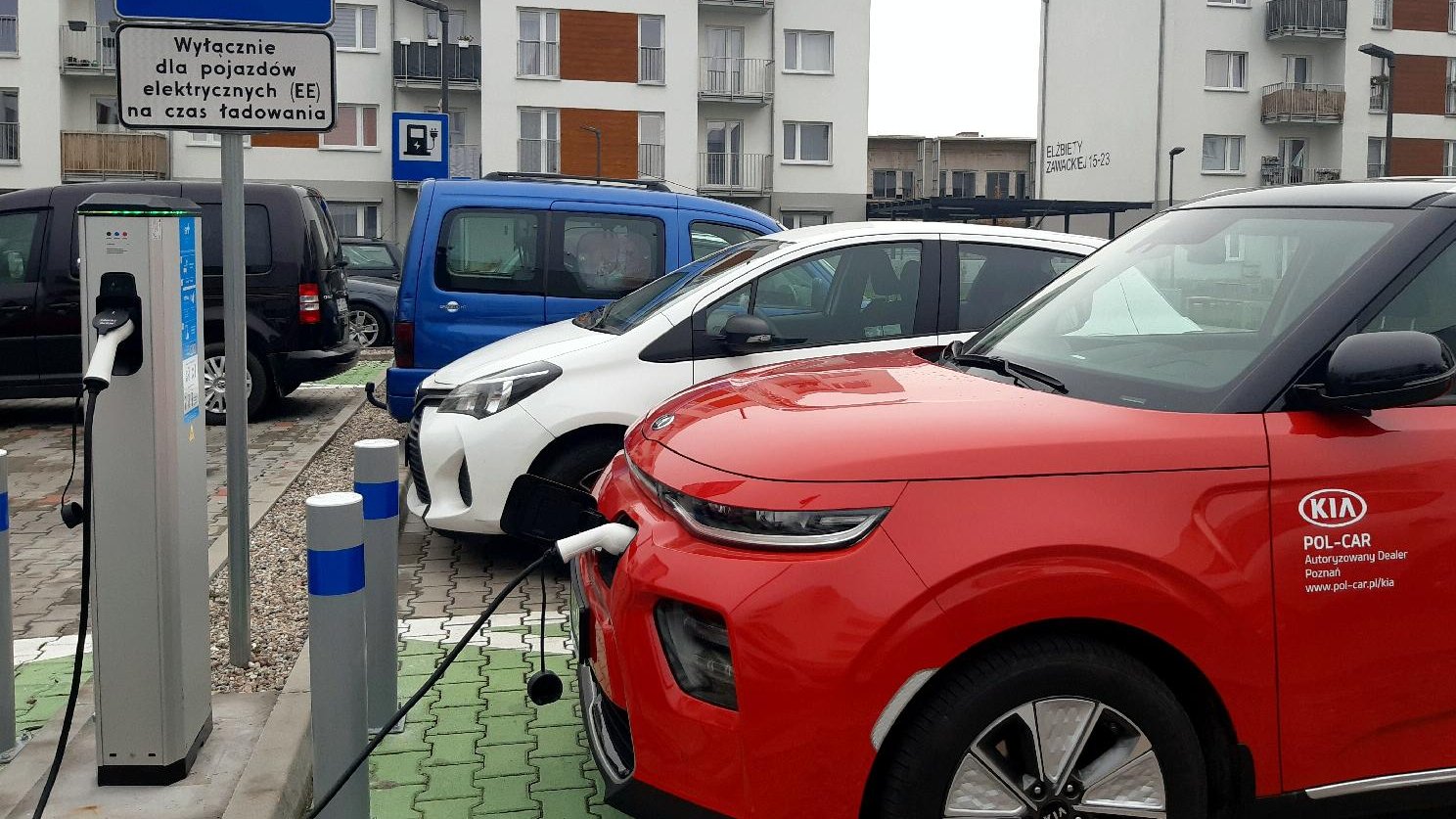 Czerwone auto podłączone kablem do stacji ładującej. W tle zabudowania mieszkalne.