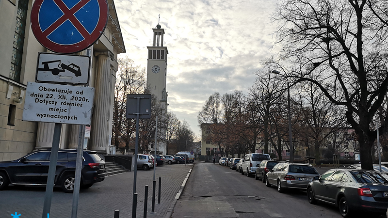 Zdjęcie przedstawia ulicę, na poboczach po obu stronach jezdni stoją zaparkowane samochody. Znajduje się też znak informujący o konieczności przeparkowania aut.