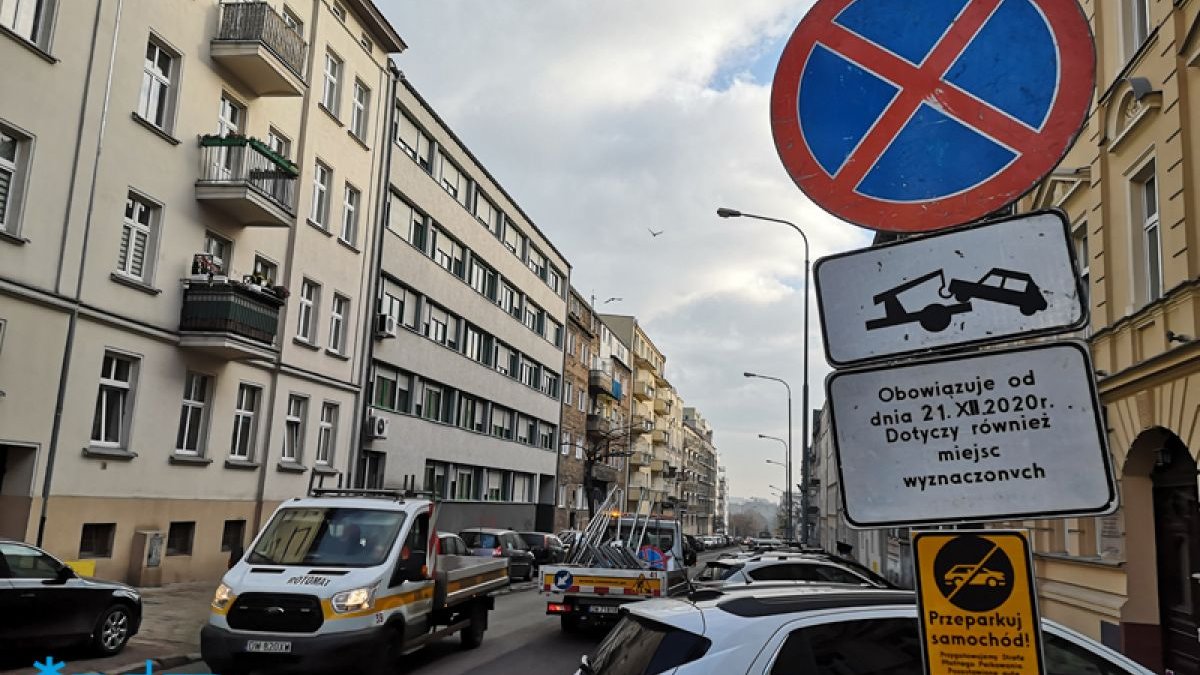 Zdjęcie przedstawia ulicę, na poboczach po obu stronach jezdni stoją zaparkowane samochody. Znajduje się też znak informujący o konieczności przeparkowania aut.