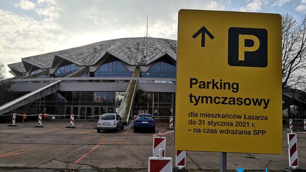 Zdjęcie przedstawia Halę Arenę przed którą stoją zaparkowane samochody. Widać też znak informujący o parkingu tymczasowym.