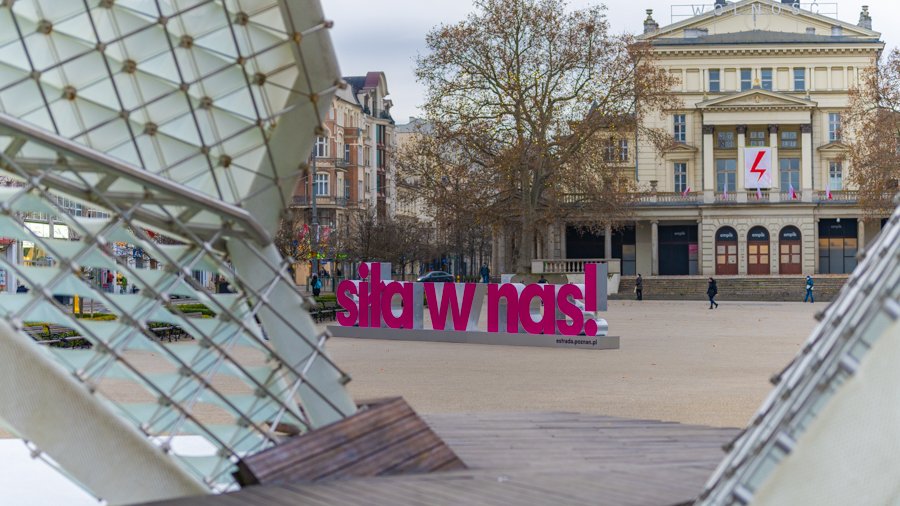 Zdjęcie przedstawia różowy neon z napisem "Siła w nas". Znajduje się on na placu Wolności. W tle widać budynek Arkadii. Na pierwszym planie widać fragment fontanny.