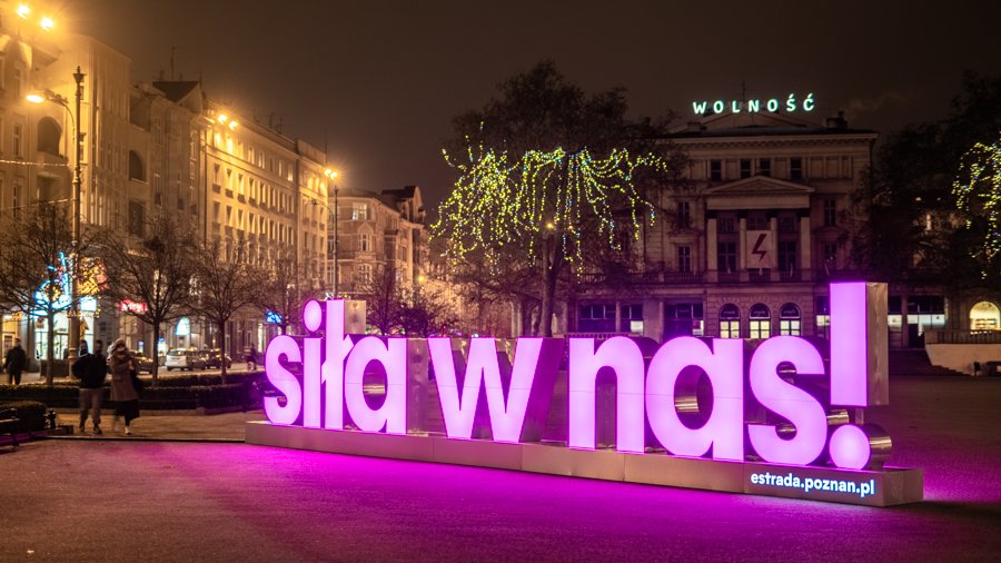 Zdjęcie przedstawia różowy, podświetlony neon z napisem "Siła w nas". Znajduje się on na placu Wolności. Zdjęcie zostało zrobione wieczorem.