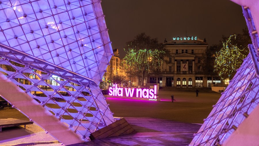 Zdjęcie przedstawia różowy, podświetlony neon z napisem "Siła w nas". Znajduje się on na placu Wolności. Na pierwszym planie widać fontannę, w tle budynek Arkadii. Zdjęcie zostało zrobione wieczorem.