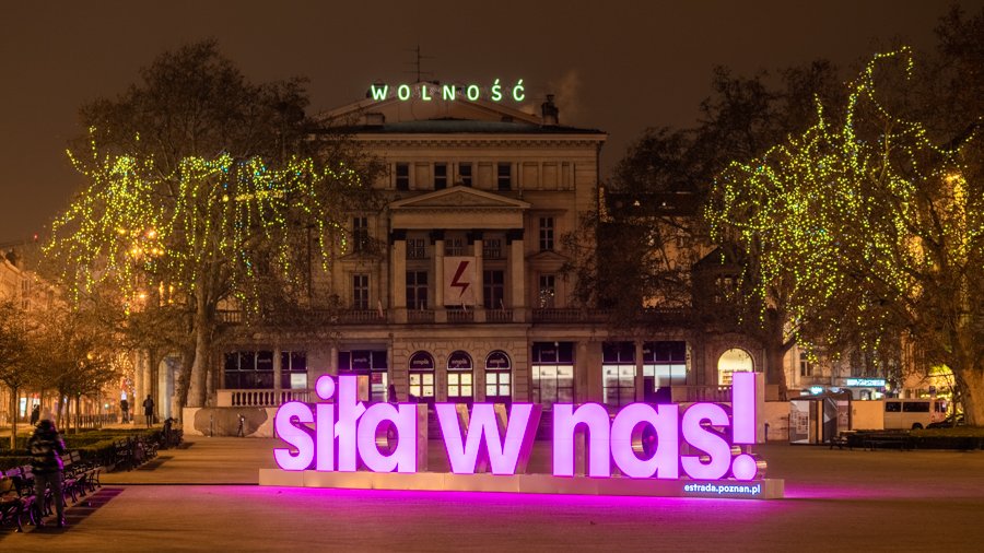 Zdjęcie przedstawia różowy, podświetlony neon z napisem "Siła w nas". Znajduje się on na placu Wolności. Zdjęcie zostało zrobione wieczorem.