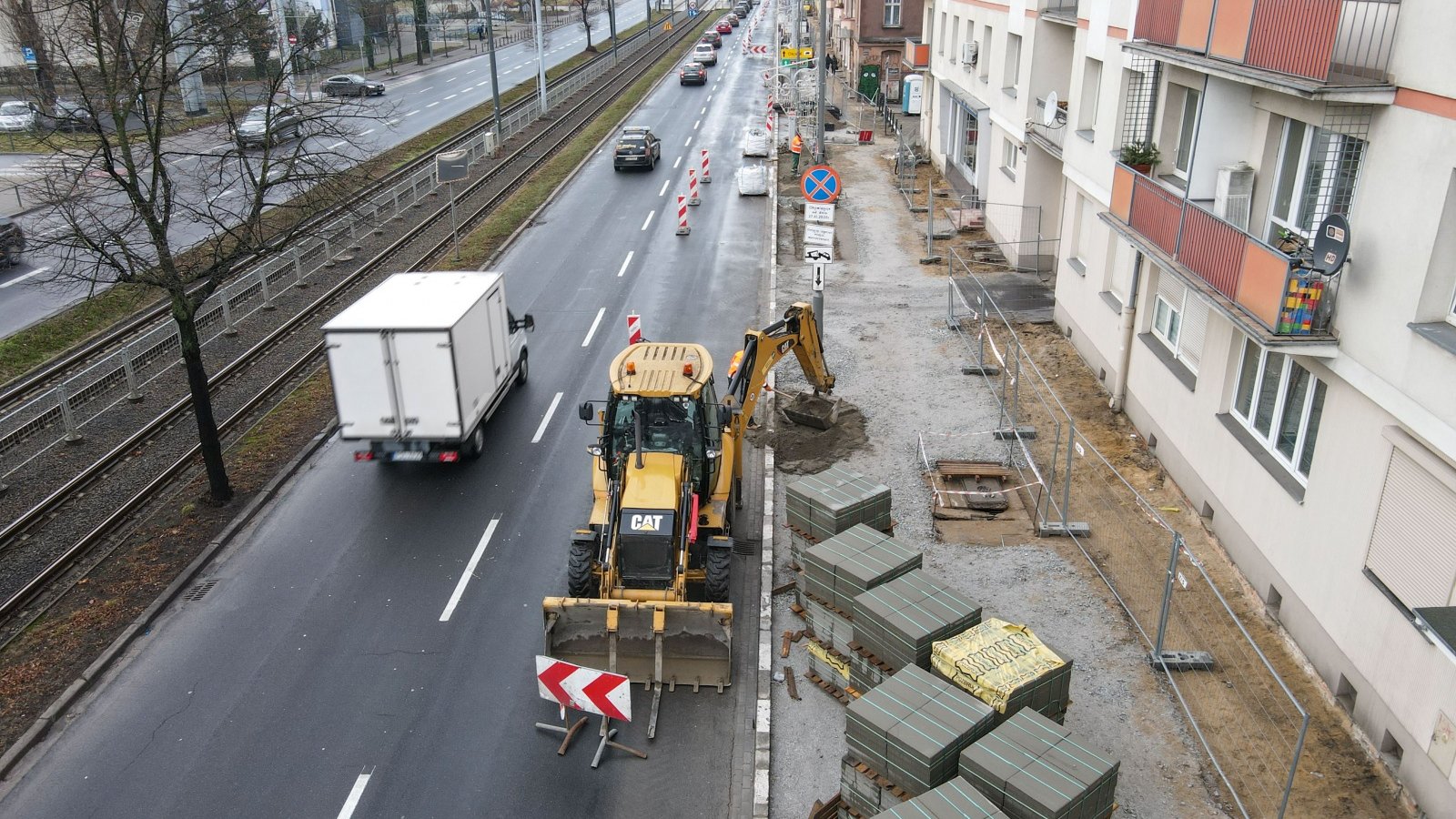 Prace nad budową nowego chodnika. Zdjęcie z góry. Widać na nim też jezdnię, samochody, maszynę budowlaną i sąsiedni budynek.