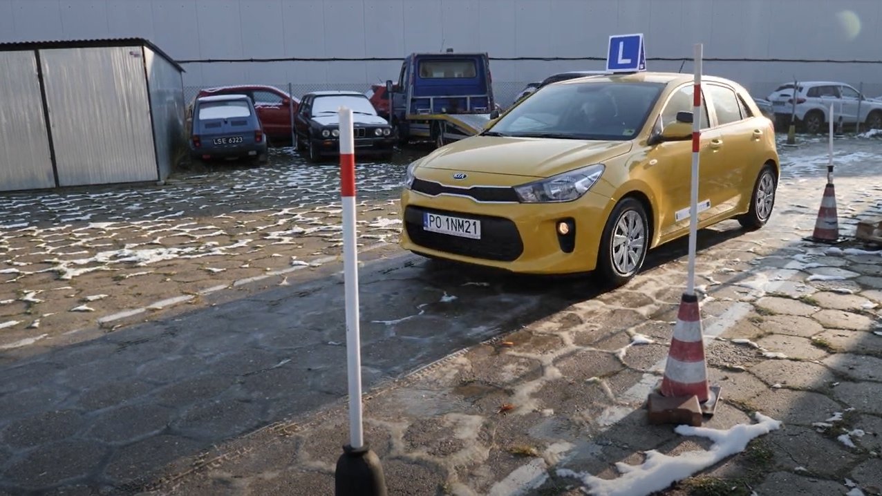 Zdjęcie przedstawia żółty samochód do nauki jazdy, tzw."elkę". Auto jedzie pomiędzy słupkami, w tle widać inne pojazdy.