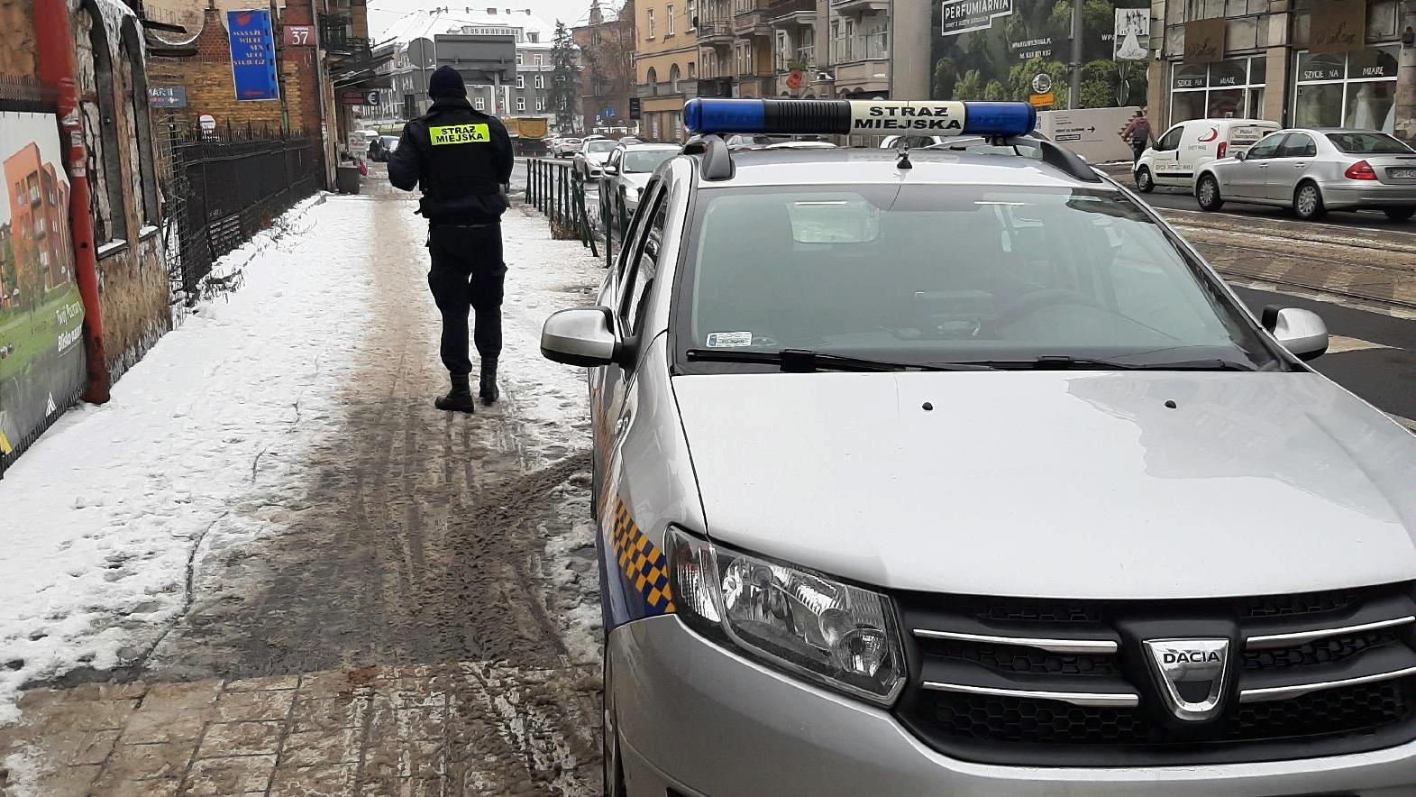 Na pierwszym planie znajduje się radiowóz straży miejskiej, stojący na poboczu przy jezdni. Na chodniku tyłem stoi strażnik miejski. W tle widać jadące po ulicy samochody oraz stojące przy drodze budynki. - grafika artykułu