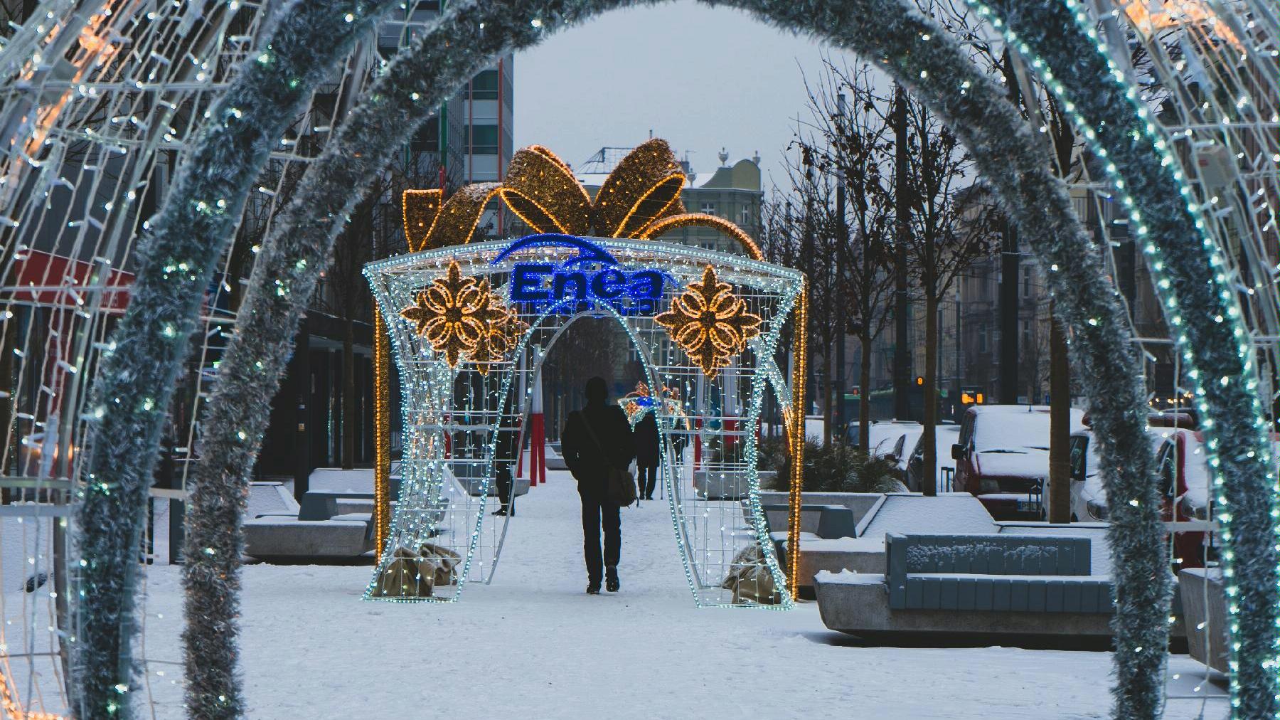 Zdjęcie przedstawia ozdoby i iluminacje świąteczne na ul. Św. Marcin. Na pierwszym planie widać świecącą dużą ozdobę w kształcie prezentu z napisem Enea, pod którą przechodzi mężczyzna w czarnym płaszczu. - grafika artykułu