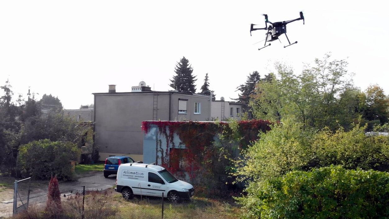 Zdjęcie przedstawia budynki jednorodzinne. Na podwórku jednego z nich, który znajduje się najbliżej stoją dwa samochody. W powietrzu unosi się dron.