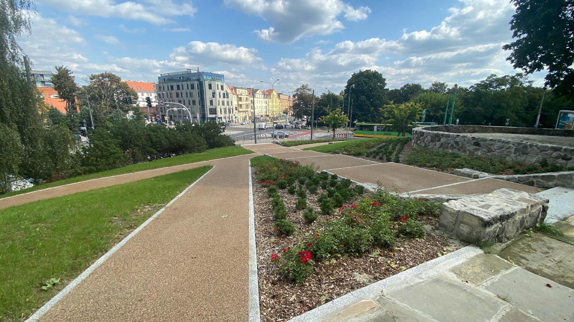 Zdjęcie przedstawia park. Widać na nim alejki spacerowe oraz roślinność. W tle znajdują się budynki oraz jezdnia, po której jadą samochody i tramwaje.