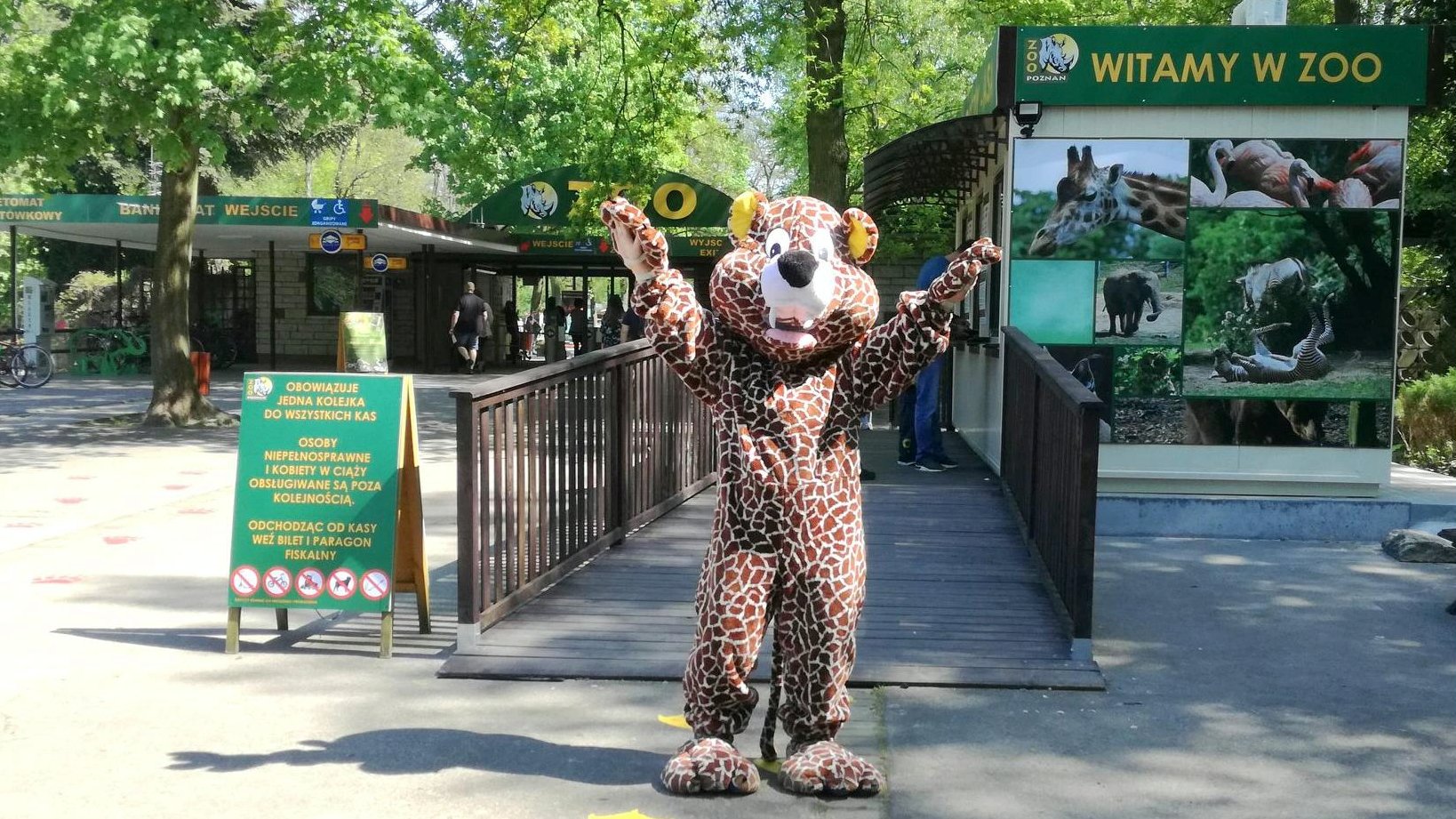 Zdjęcie przedstawia wejście do zoo. Na pierwszym planie widać osobę przebraną za geparda - stoi ona z otwartymi ramionami, w geście przywitania. W tle widać kasy, bramki wejściowe i napis "witamy w zoo". - grafika artykułu