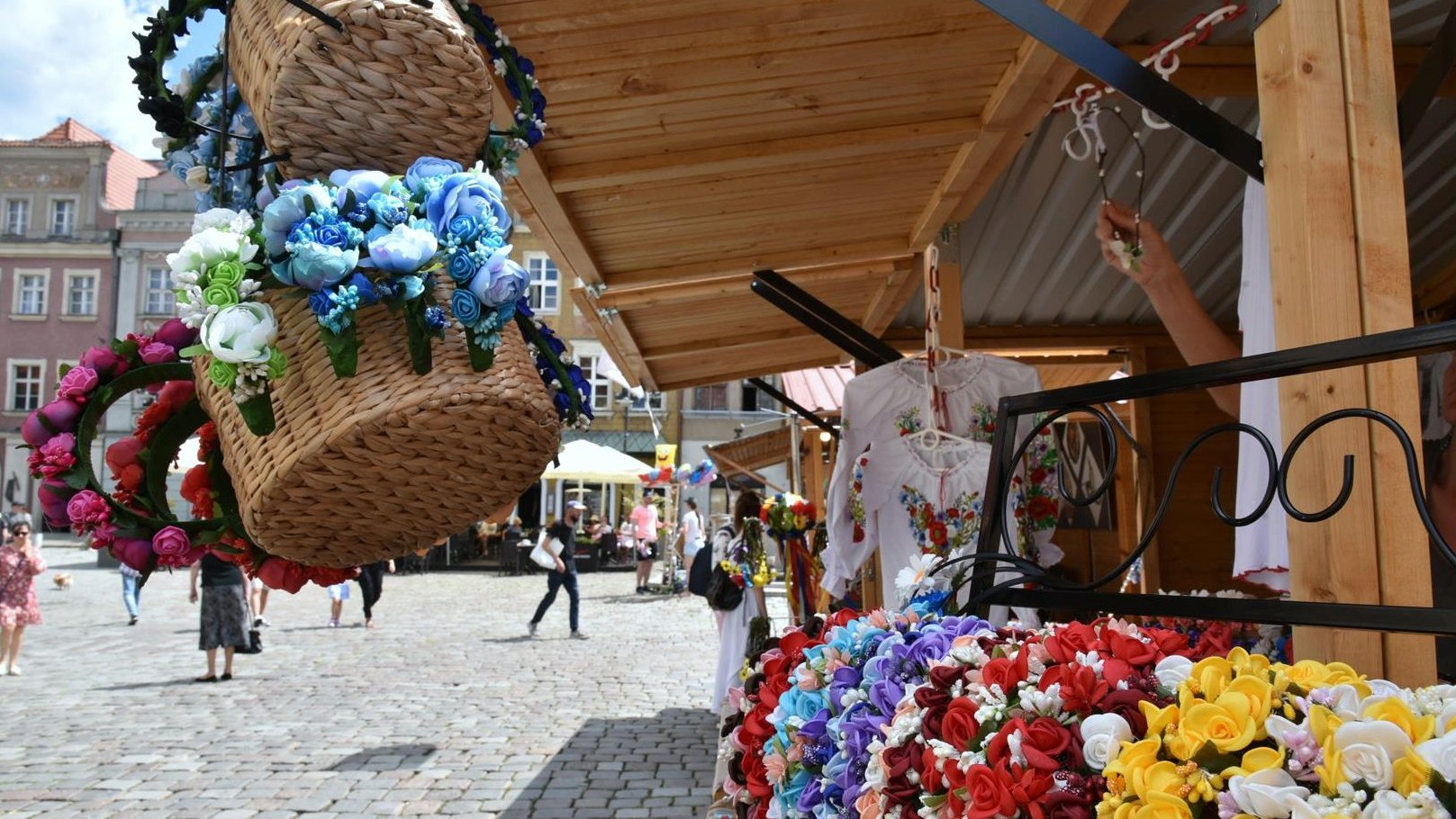 Zdjęcie przedstawia drewniany domek handlowy stojący na Starym Rynku. Na stoisku znajdują się bukiety ze sztucznych kwiatów (niektóre umieszczone zostały w wiklinowych koszykach) oraz bluzki w kwiatki.