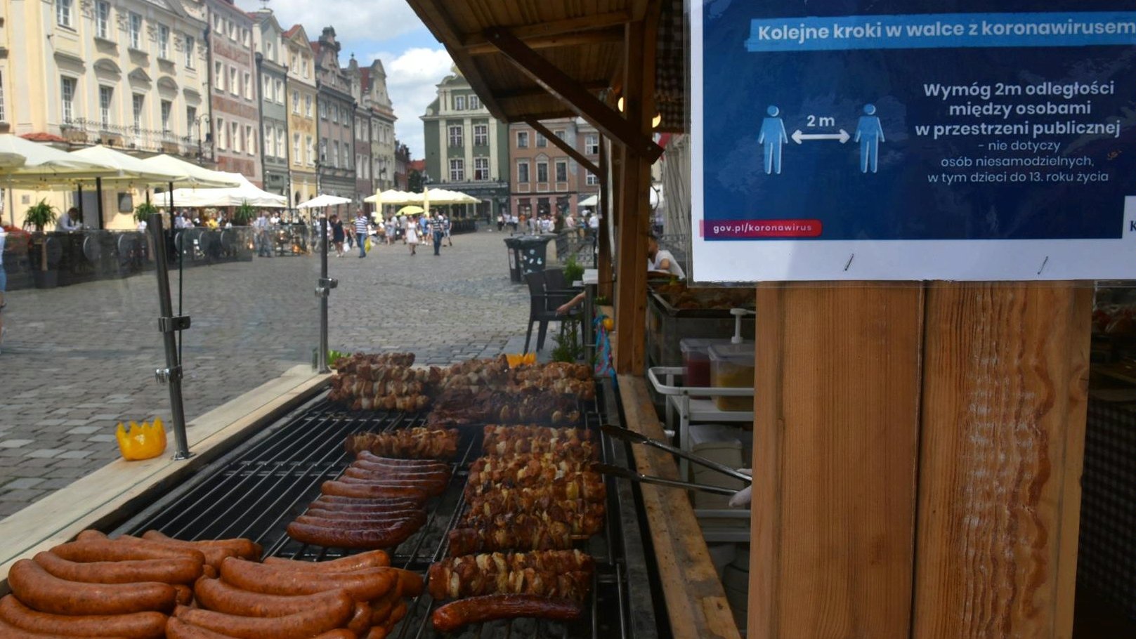 Zdjęcie przedstawia stoisko z kiełbasami i szaszłykami z grilla. Na pierwszym planie widać plakat informujący o zasadach bezpieczeństwa, jakich należy przestrzegać w związku z covid-19.