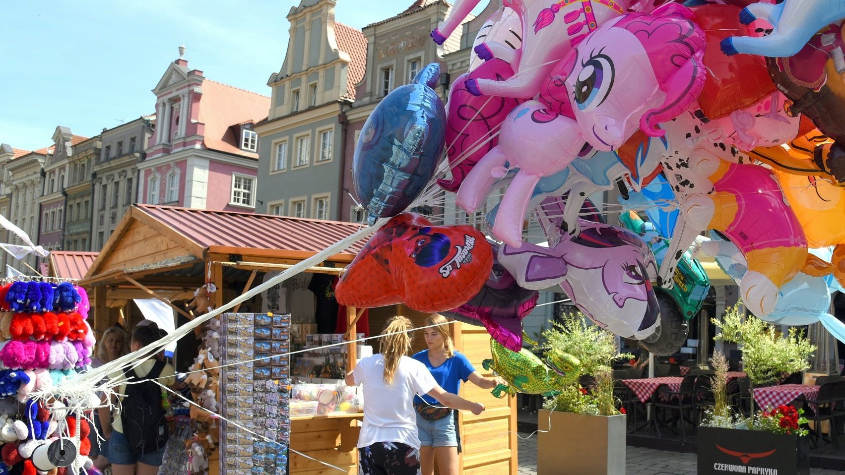 Zdjęcie przedstawia drewniany domek, na którym sprzedawane są pamiątki związane z Poznaniem, m.in. pluszowe koziołki czy magnesy. Na pierwszym planie znajdują się balony na sznurkach.