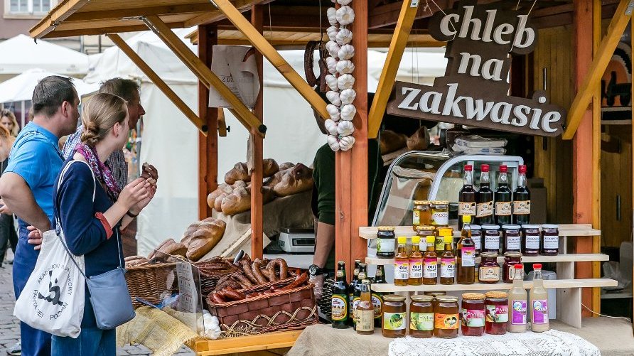 Zdjęcie przedstawia drewniane stoisko, na którym sprzedawane są chleb, wędliny, kwas chlebowy, miód, oleje i soki owocowe. Widać tez duży napis "chleb na zakwasie". Przy stoisku stoją ludzie.