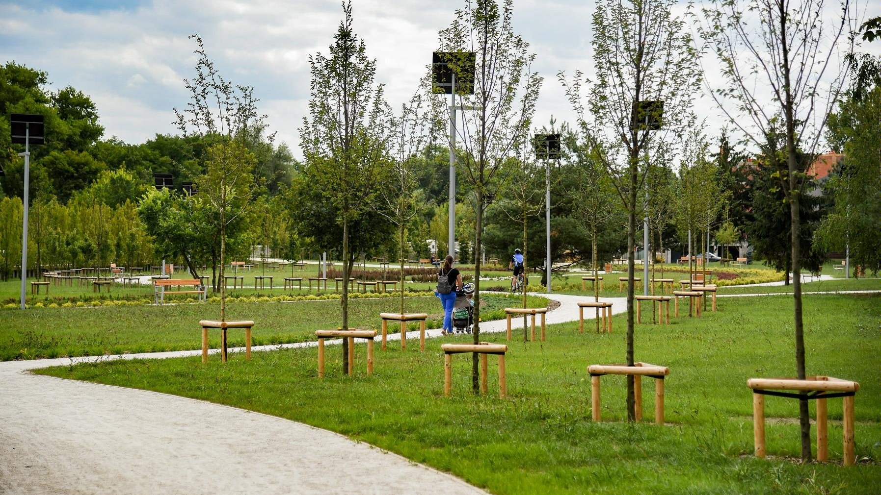 Zdjęcie przedstawia teren zielony - trawę, drzewa, krzewy. Przez park ciągną się alejki spacerowe, po których chodzą ludzie, np. matka z dzieckiem w wózku. Jeżdżą po nich także rowerzyści. Obok stoją ławki.