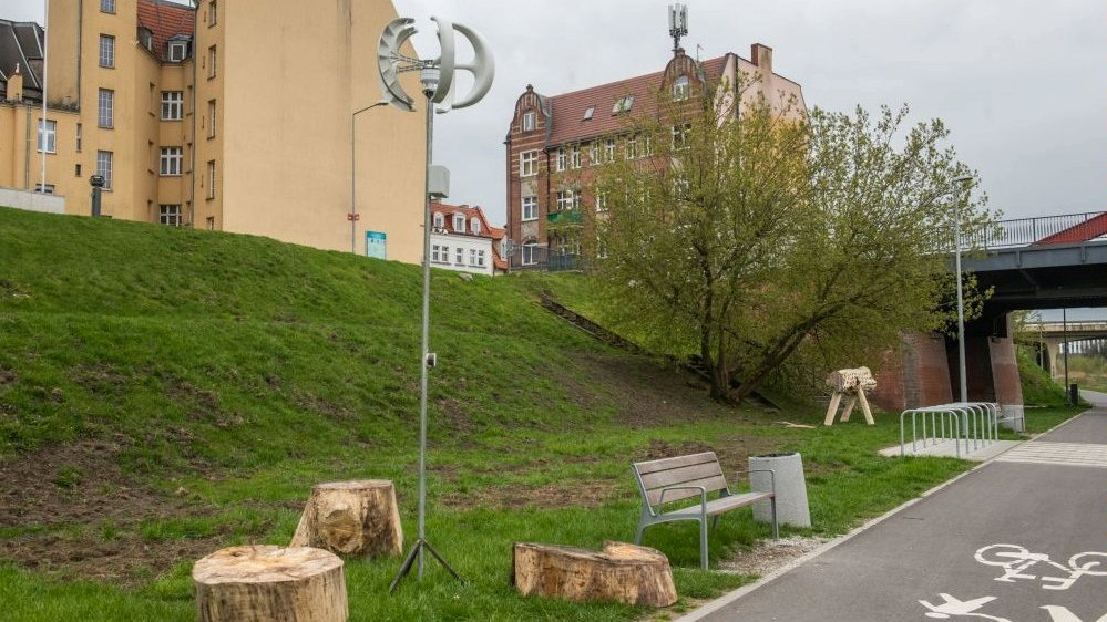 Zdjęcie przedstawia drewniane pieński i instalację pozwalającą na ładowanie urządzeń dzięki sile wiatru - słup zakończony wiatrakiem. Obok znajduje się droga rowerowa. Z tyłu widać budynki.