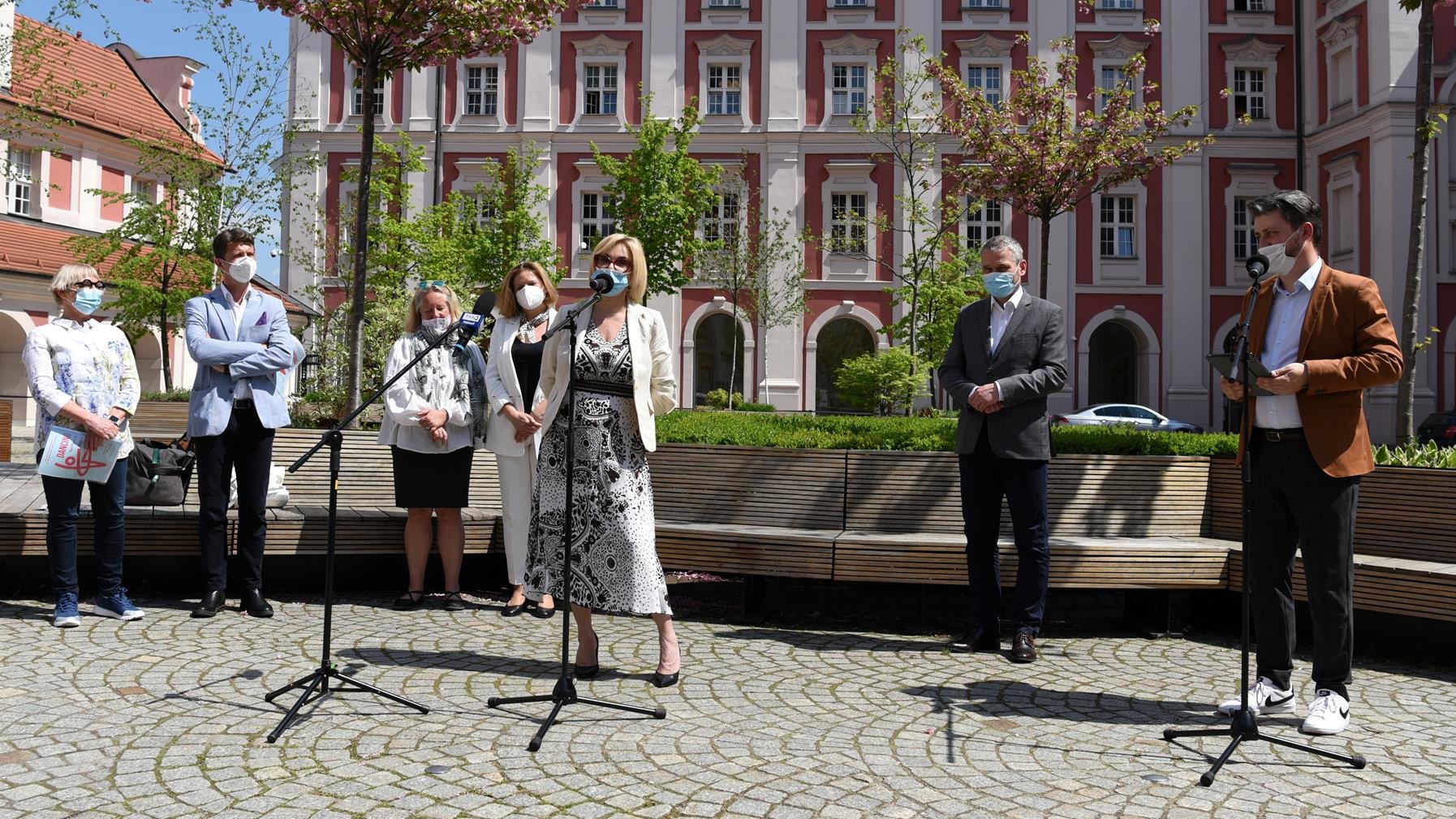 Zdjęcie z konferencji prasowej. Na pierwszym planie jej uczestnicy, w tle budynek urzędu miasta na placu Kolegiackim