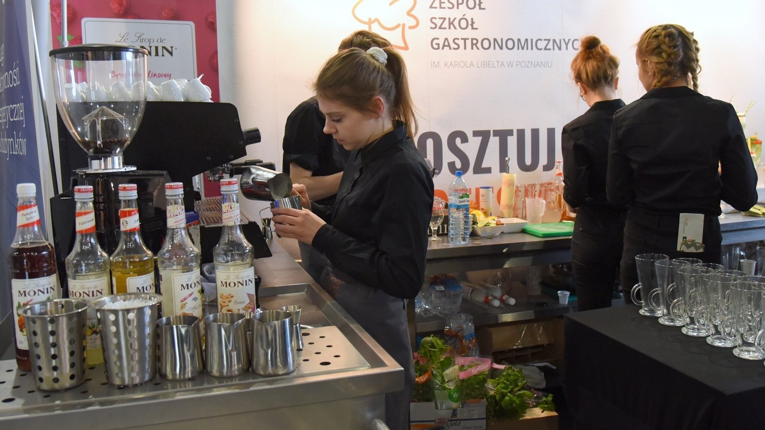 Zdjęcie przedstawia dziewczyny w czarnych koszulach na stanowisku gastronomicznym. Widać na nim takie akcesoria, jak szklanki, ekspres do kawy czy smakowe syropy do kawy w szklanych butelkach.