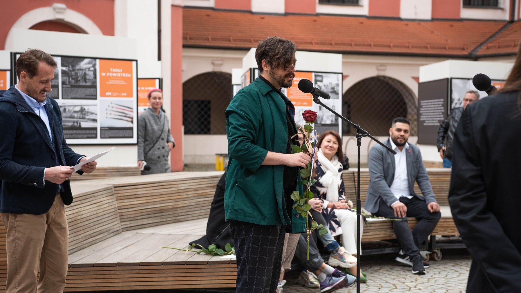 Galeria zdjęć z uroczystości wręczenia stypendiów - przemawia jeden z laureatów, w ręce trzyma czerwoną róże, w tle widać innych ludzi, w tym nagrodzonych laureatów.