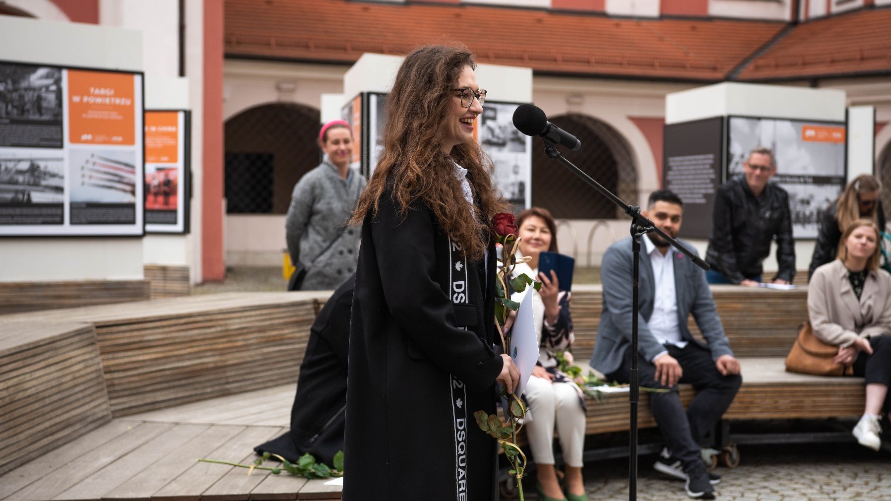 Galeria zdjęć z uroczystości wręczenia stypendiów - przy mikrofonie jedna z laureatek, w tle widać pozostałych nagrodzonych siedzących na ławce.