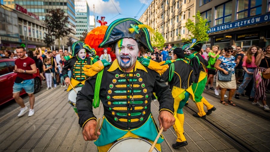 Parada na ul. Św. Marcin. W środku kadru przebrany w zielono-czarny mundur artysta, który gra na werblu. W tle pozostali członkowie trupy artystycznej oraz widzowie.