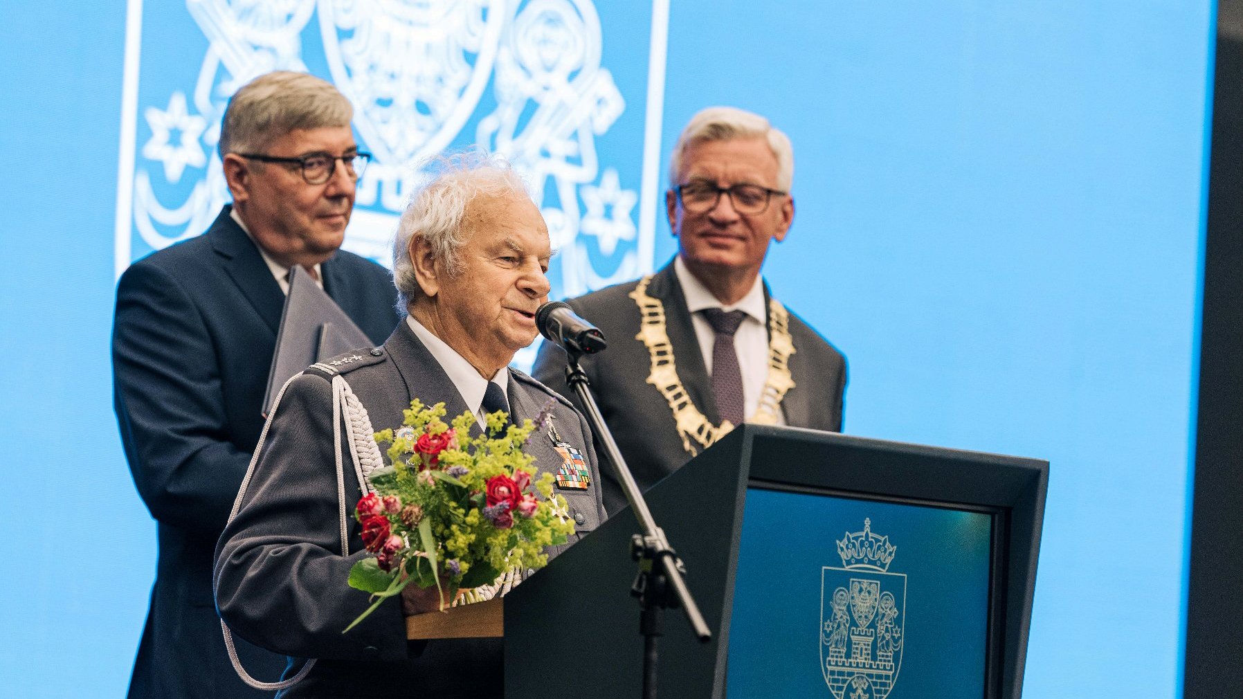 Hieronim Kowalski przy mównicy, trzyma kwiaty. Za nim Grzegorz Ganowicz i Jacek Jaśkowiak