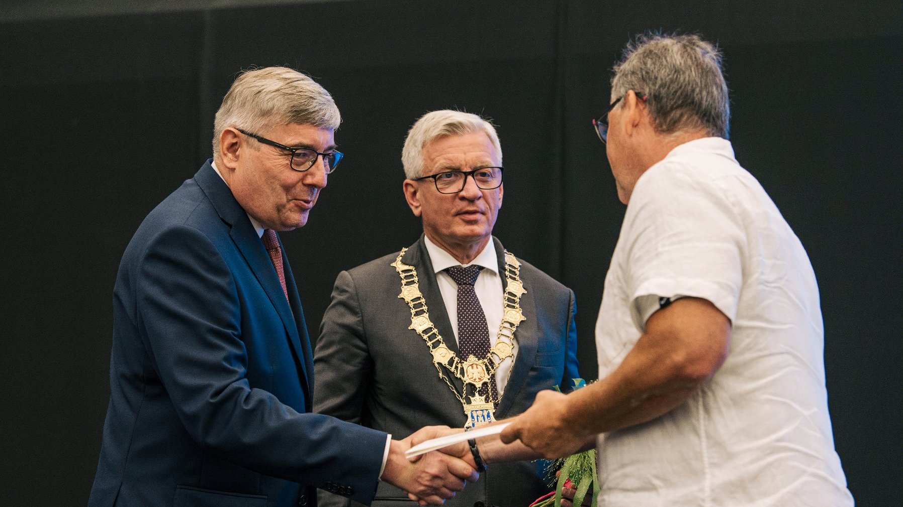 Grzegorz Ganowicz i JAcek Jaśkowiak przekazują nagrodę stojącemu tyłem do obiektywu mężczyźnie w białej koszuli