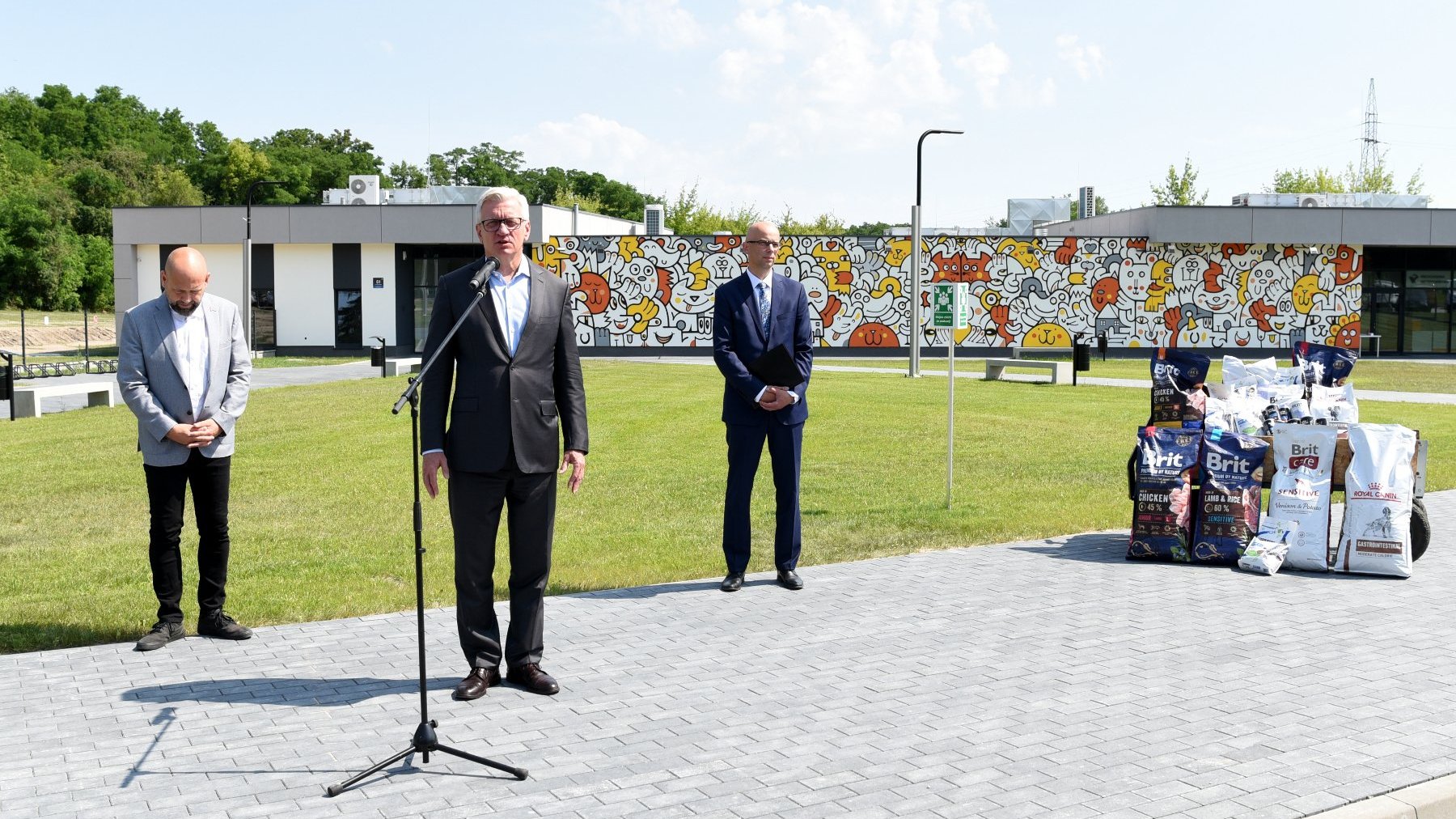 Trzej mężczyźni stoją przed mikrofonami, w tle budynek schroniska, obok torby z karmą dla zwierząt