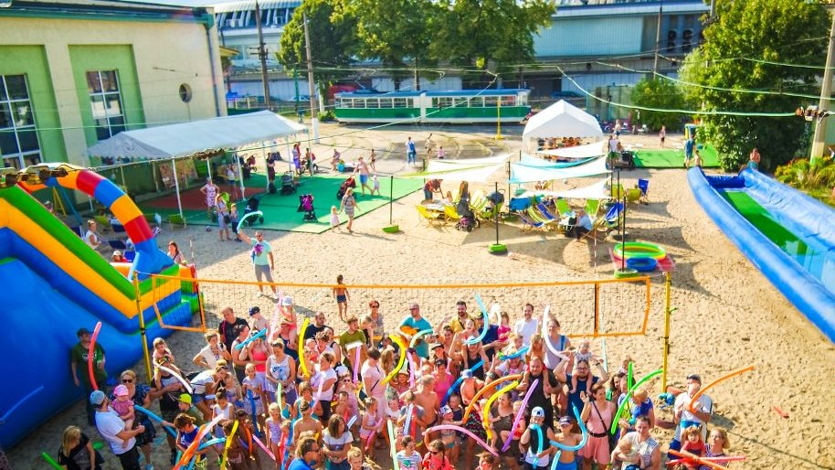 Zdjęcie przedstawia dawną zajezdnie tramwajową, na której znajduje się plaża, tor wodny, dmuchane atrakcje czy boisko do siatkówki. Na zdjęciu widzimy tłum ludzi.