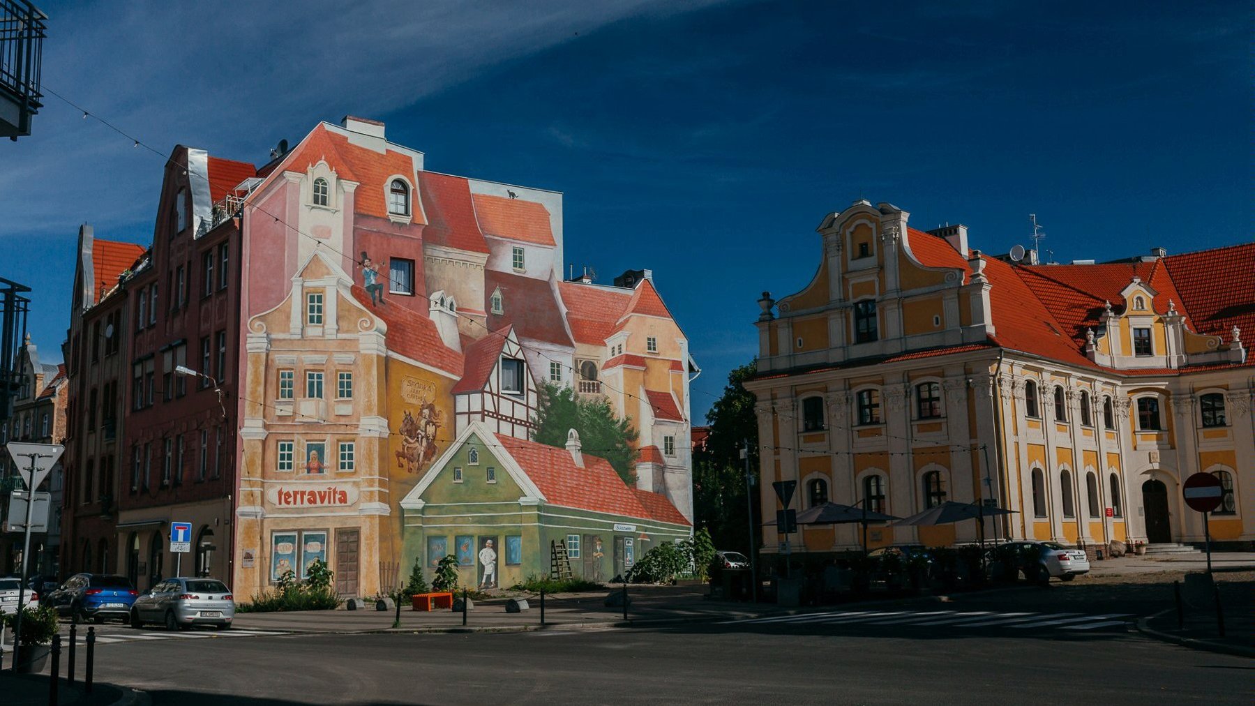 Zdjęcie przedstawia mural. Obraz na ścianie obrazuje budynki i robi wrażenie malowidła 3D.