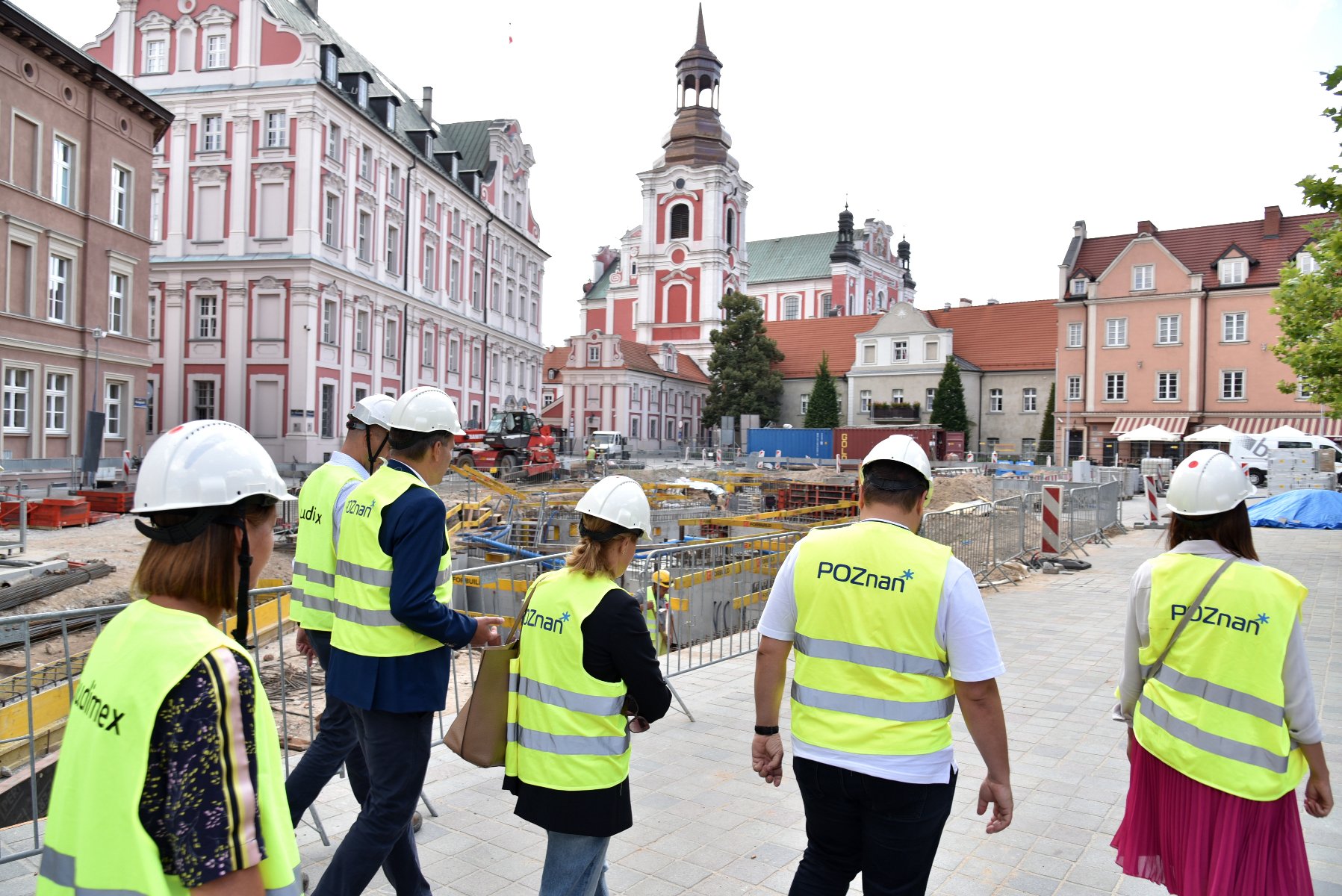 Plac Kolegiacki. Na pierwszym planie grupa osób w kaskach i kamizelkach odblaskowych. Na dalszym planie plac budowy. W tle budynek urzędu miasta. - grafika artykułu