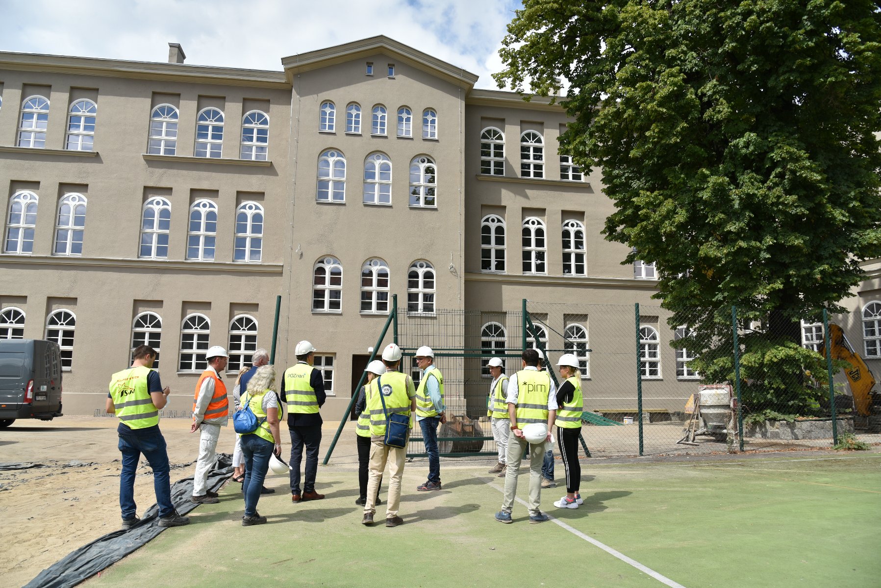 Zdjęcie przedstawia grupę ludzi w żółtych kamizelkach stojących przed remontowanym budynkiem szkoły. - grafika artykułu