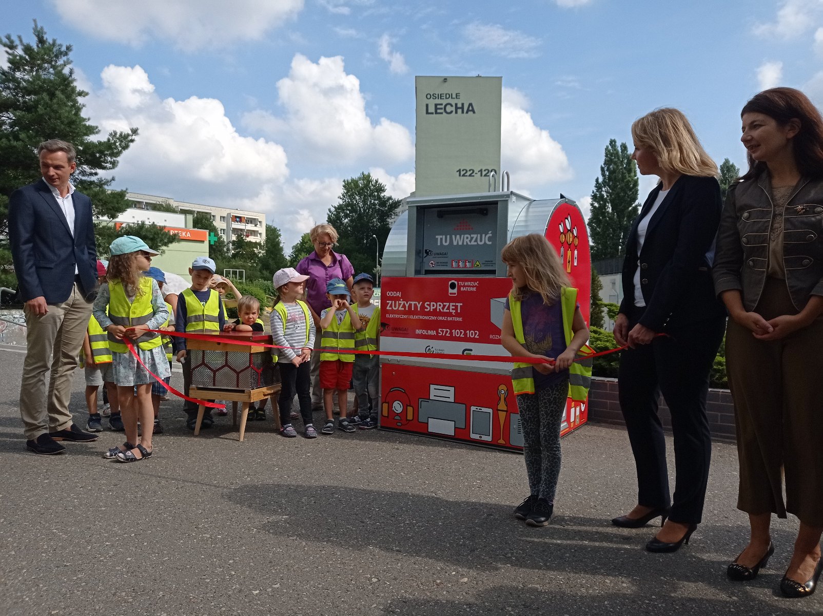 Zdjęcie przedstawia czerwony pojemnik na elektroodpady. Obok stoją dzieci w żółtych kamizelkach i ich opiekunowie. - grafika artykułu